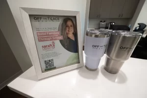 Framed promotional material for Off The Trace Dental featuring a smiling woman, alongside two branded tumblers, emphasizing family and dental care.